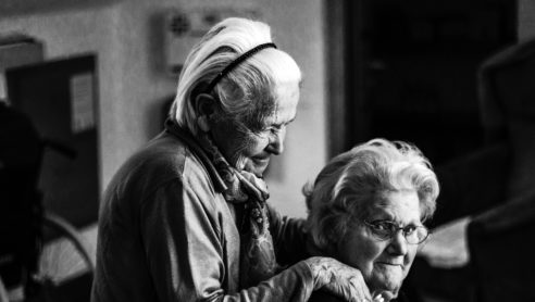 Äldre kvinna står bakom och håller om annan äldre kvinna.